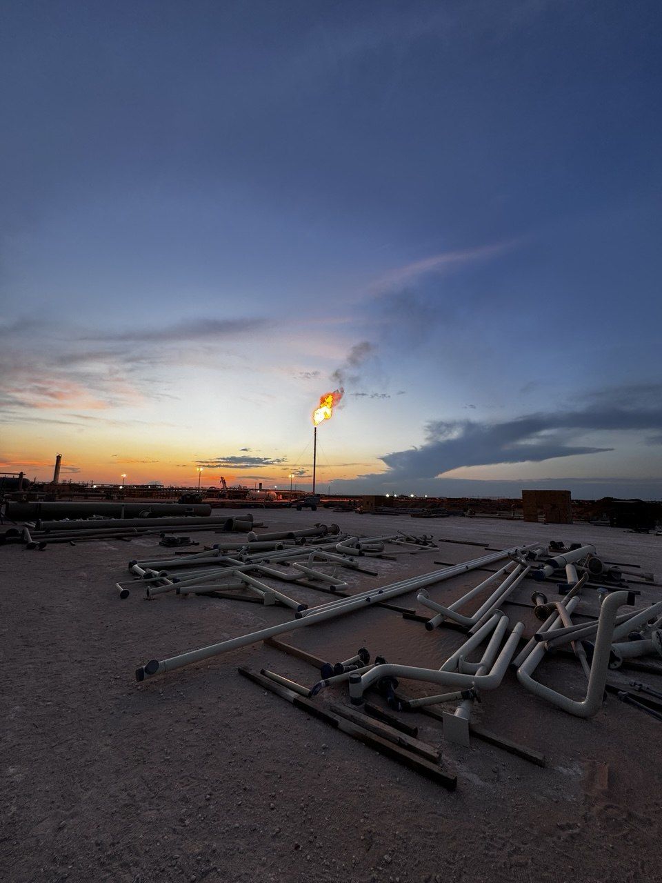 Flare stack burning at dusk above fabricated pipe spools and fittings laid out in a yard