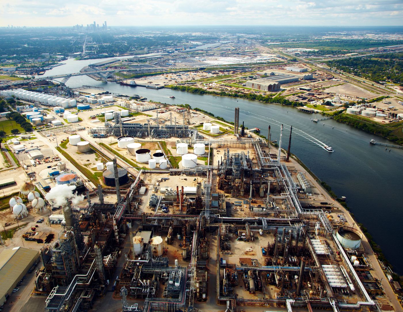 Aerial view of refinery process units and storage tanks along a ship channel, city skyline on the horizon