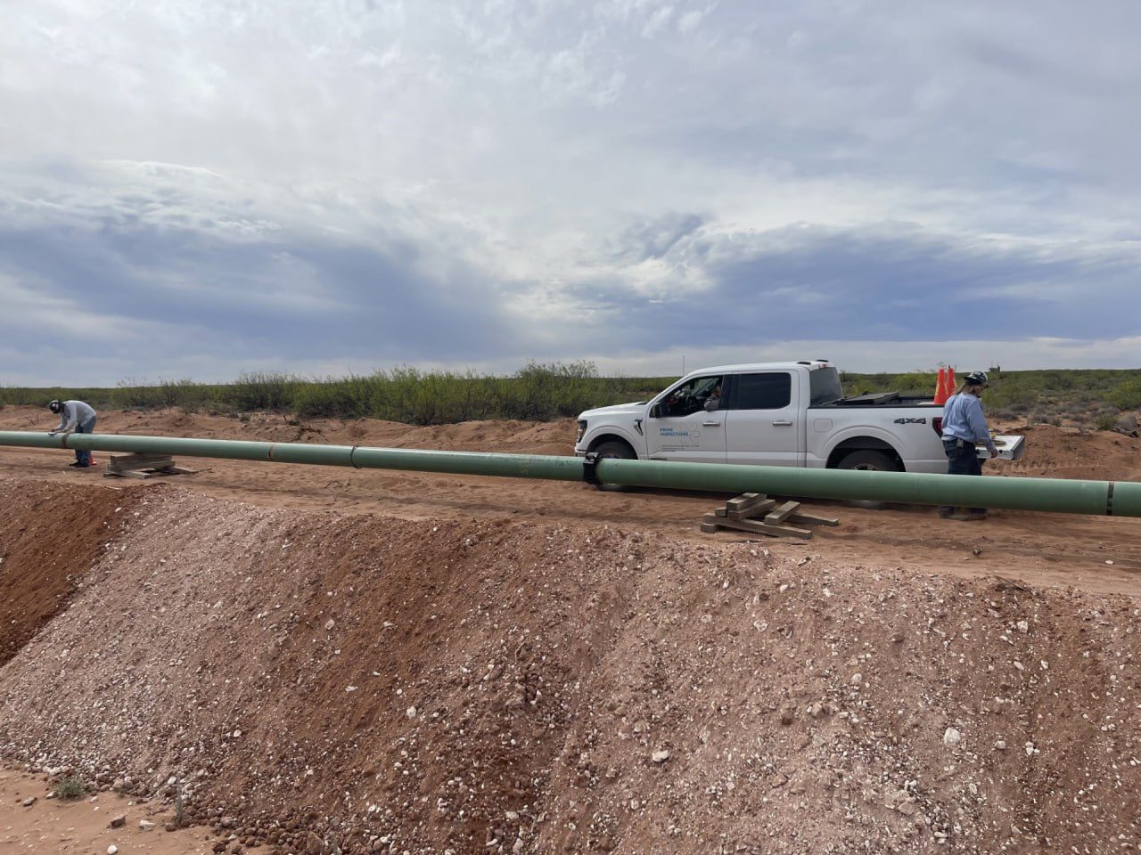 Prime Inspections truck on a pipeline right-of-way beside a green-coated pipe string, with crew working at the joints