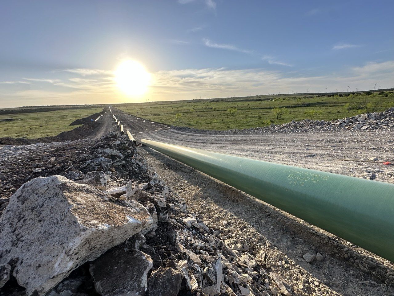 Pipeline string running toward the sunrise across open rangeland, with wind turbines on the horizon