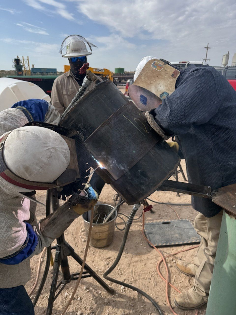 Two welders making a qualification test weld on a pipe coupon while an inspector in a hard hat and face shield watches